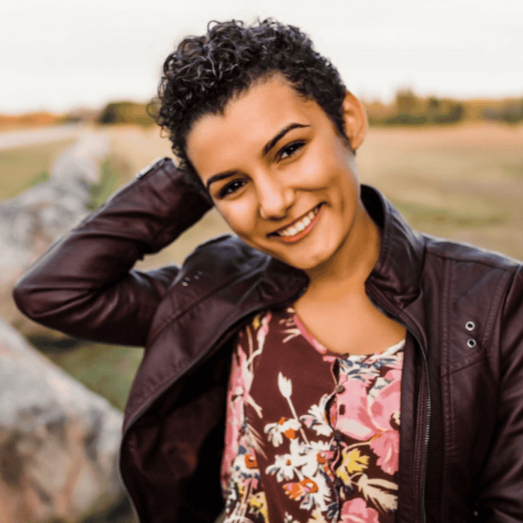 Person with short curly hair smiling, wearing a floral top and a leather jacket, stands outdoors with one hand behind their head, embodying the carefree spirit that defines ZIPDRIP's vibrant style.