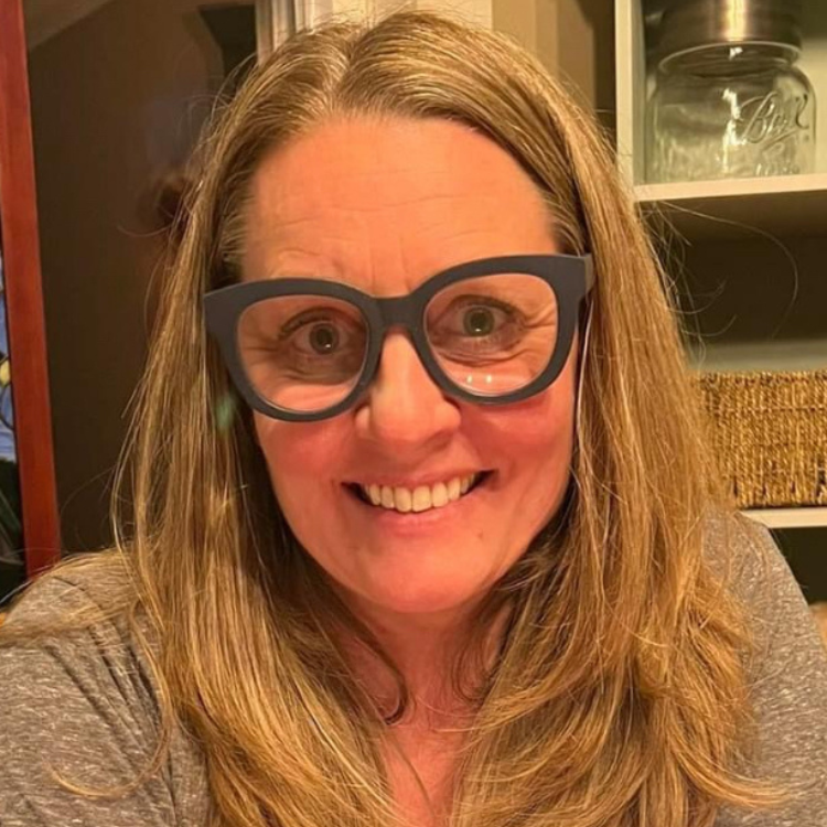 A person with long hair and large glasses is smiling at the camera in a room with shelves, curious about ZIPDRIP.