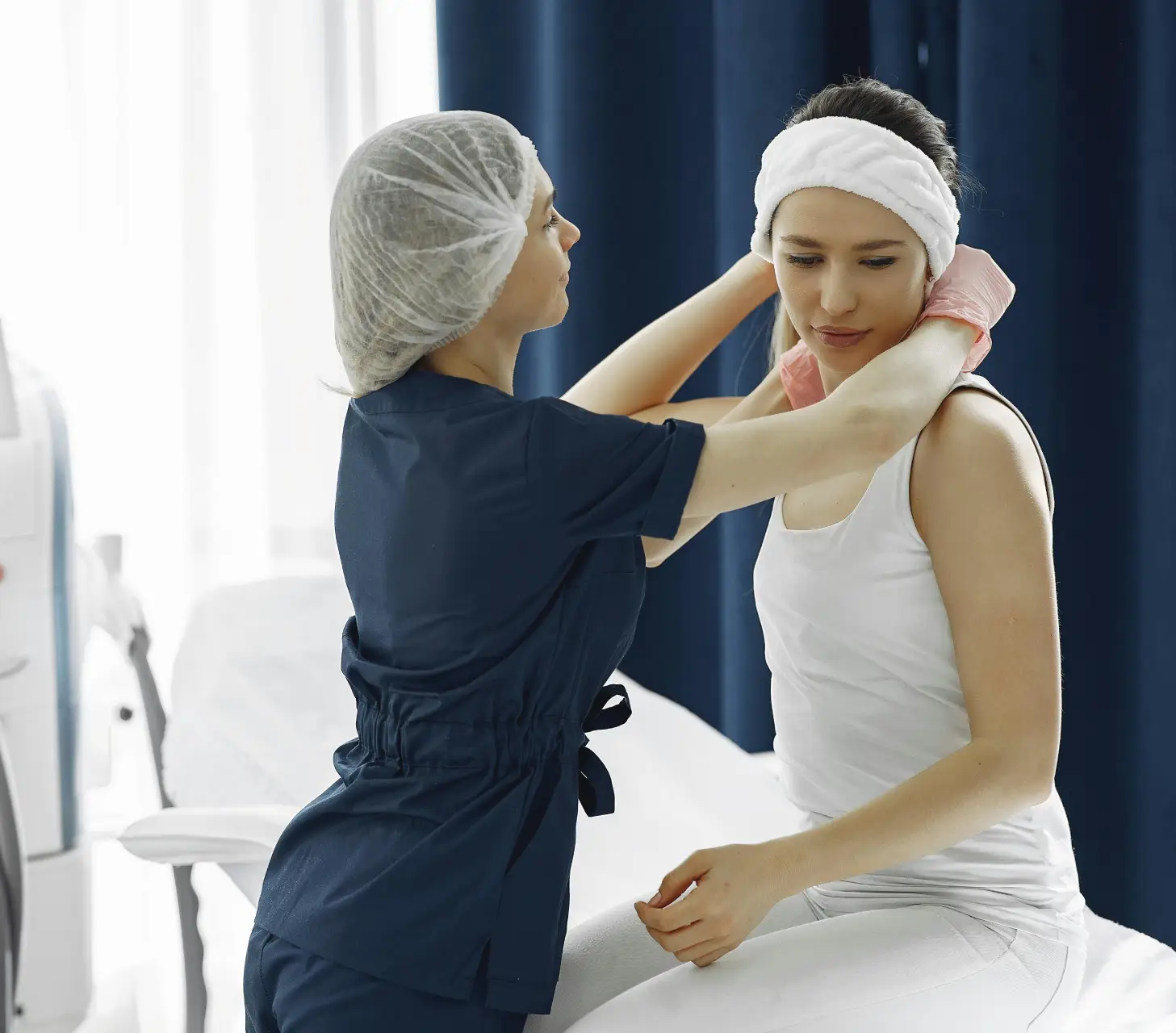 A healthcare worker in blue scrubs adjusts a white headband on a seated woman in a white tank top, preparing for mobile IV therapy in a clinical setting with dark curtains.