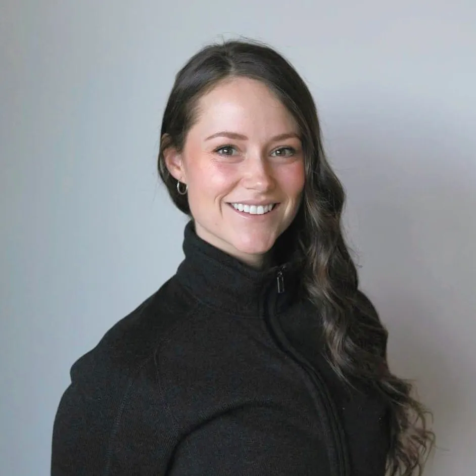 Person with long, wavy hair smiles, wearing a black zip-up jacket against a plain background.