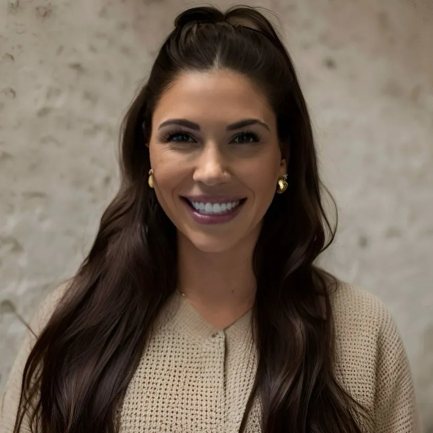 Smiling woman with long brown hair and a beige sweater against a textured wall.