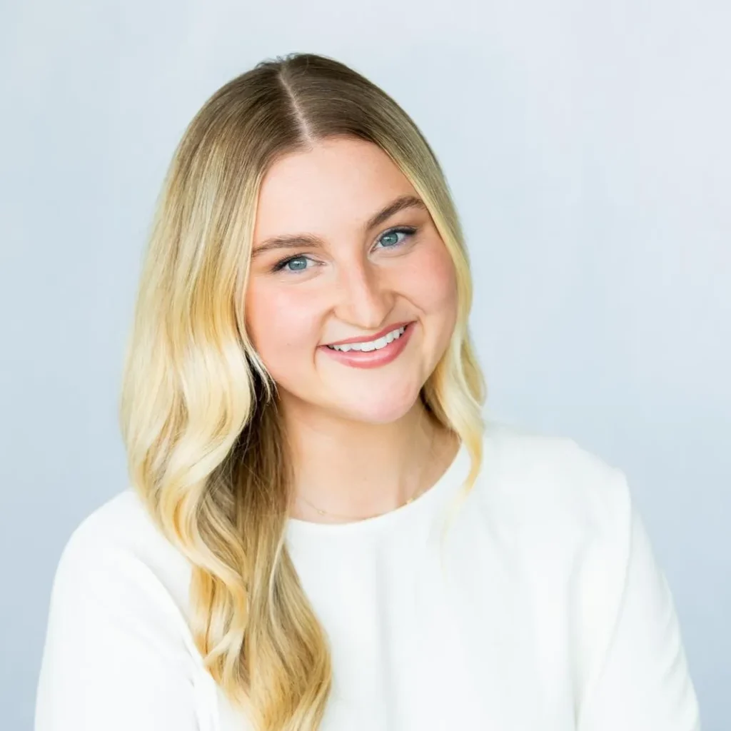 A woman with long blonde hair and a white top smiles against a light background.