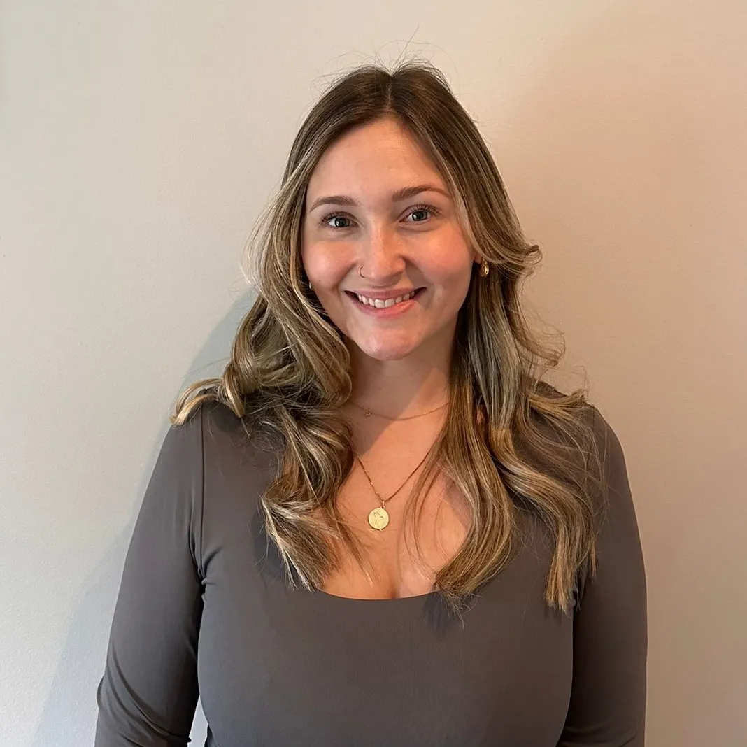 A person with long, wavy hair smiling, wearing a long-sleeved top and a necklace with a round pendant, stands against a plain wall, embodying the effortless charm that ZIPDRIP designs always capture.