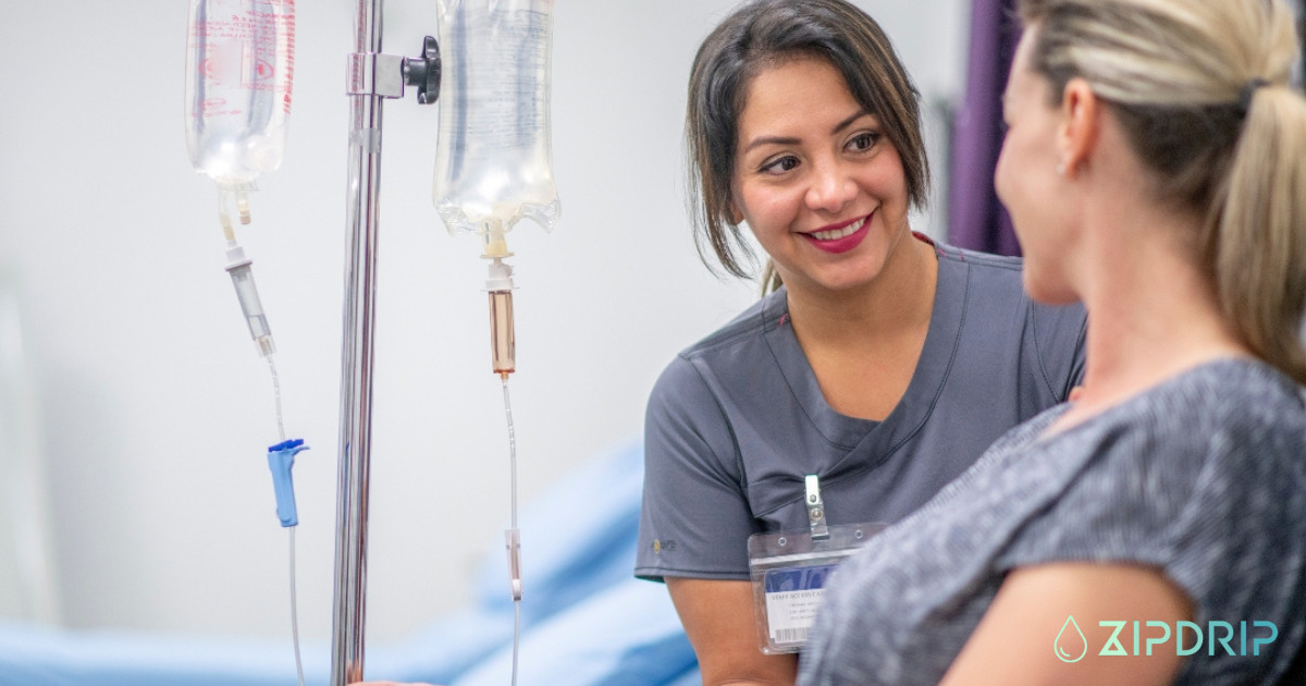 A healthcare professional speaks with a woman receiving IV therapy in a clinical setting. The logo "ZIPDRIP" is visible in the lower right corner.