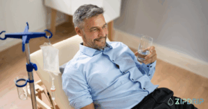 A man in a button-down shirt reclines on a chair, holding a glass of water with an IV drip stand beside him. The ZIPDRIP logo appears in the bottom right corner.