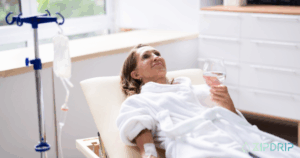 A woman in a white robe reclines on a lounge chair with an IV in her arm, holding a glass of water. An IV drip stand is beside her. The scene appears to be in a clinic or spa.