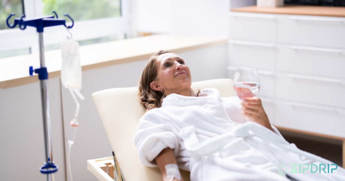 A woman in a white robe reclines on a lounge chair with an IV in her arm, holding a glass of water. An IV drip stand is beside her. The scene appears to be in a clinic or spa.