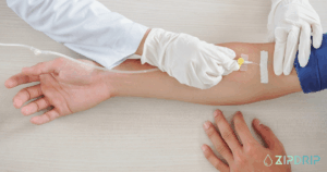 A healthcare professional wearing gloves inserts an IV drip into a person's arm, with medical tape securing the needle. The ZipDrip logo is visible in the bottom right corner.