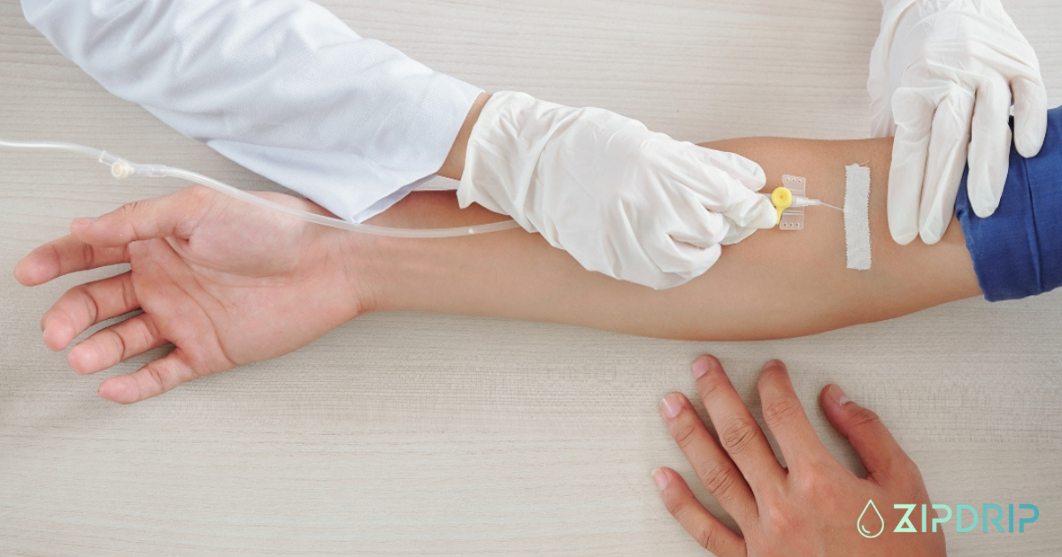A healthcare professional wearing gloves inserts an IV drip into a person's arm, with medical tape securing the needle. The ZipDrip logo is visible in the bottom right corner.
