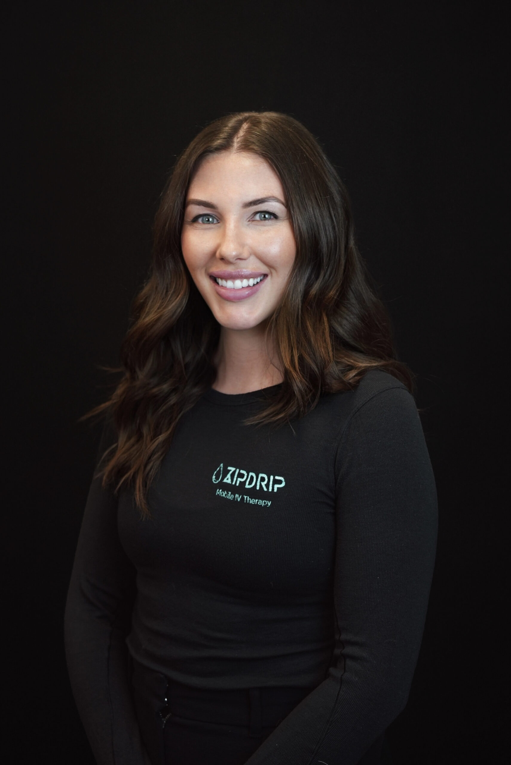 A woman with long brown hair and a black long-sleeve shirt that says "JP DRIP Mobile IV Therapy" smiles in front of a plain black background, showcasing her enthusiasm for About ZIPDRIP and its mobile wellness services.