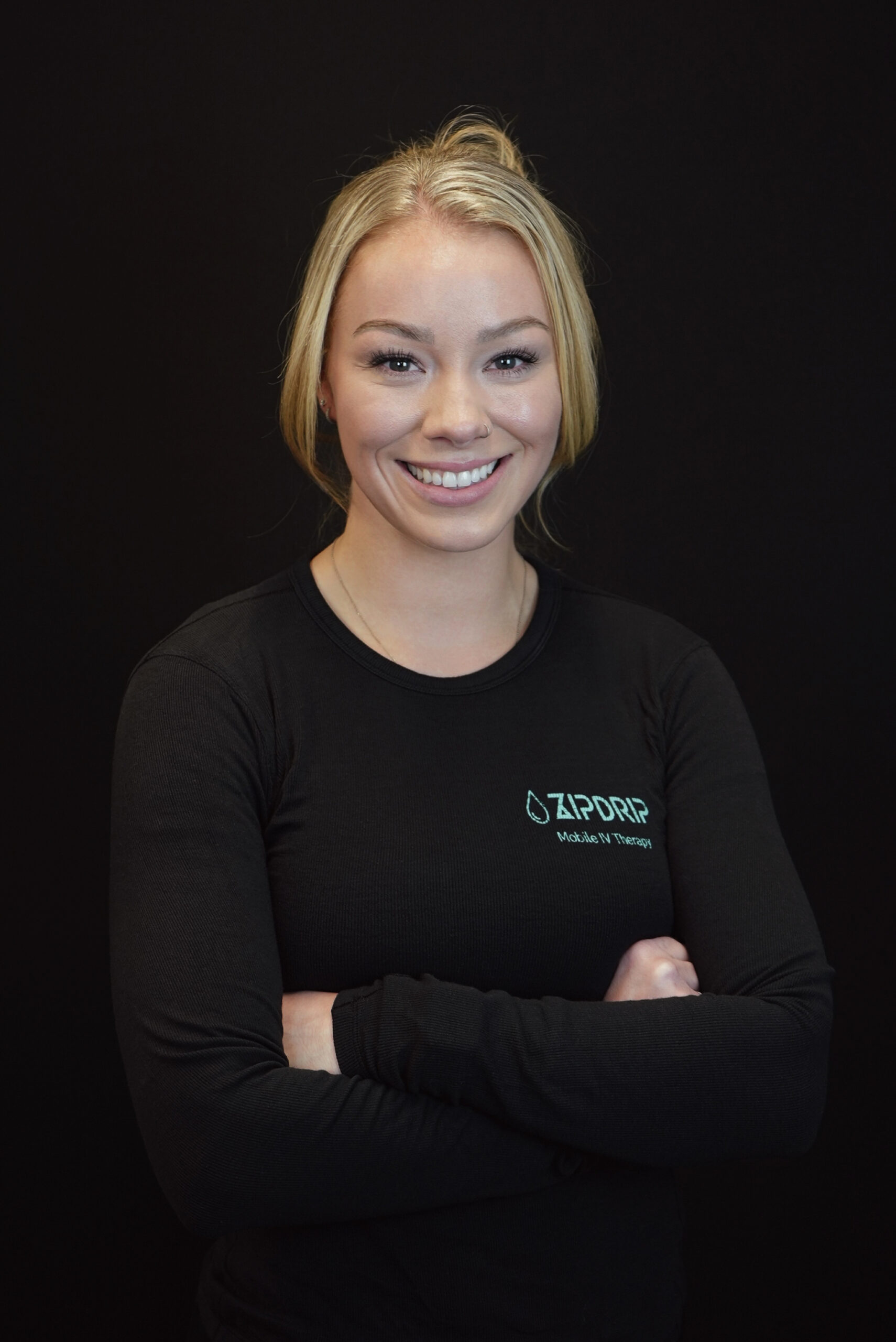 A woman with blonde hair smiles at the camera with arms crossed, wearing a black long-sleeve "ZIPDRIP Mobile IV Therapy" shirt against a plain black background, embodying the professionalism and care central to About ZIPDRIP.