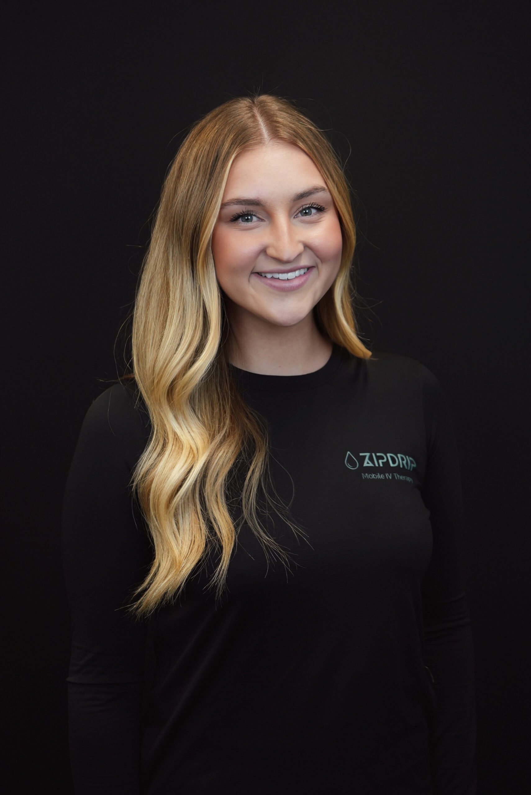 A woman with long, wavy blonde hair wearing a black shirt with the ZapDRIP logo stands against a plain black background, smiling at the camera—capturing the spirit of About ZIPDRIP.