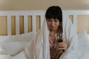 A woman sits on a bed wrapped in a white blanket, holding a glass of red wine and appearing upset—could she be wondering, Can Mobile IV Therapy Help with a Hangover?.