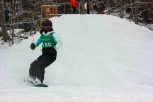 A snowboarder in a green vest and helmet rides down a snowy slope in a wooded area, while people stand at the top—showcasing the kind of winter activity where Mobile IV immune support can benefit athletes in Vancouver.