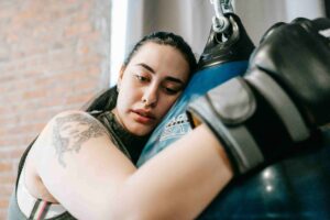Woman wearing boxing gloves rests her head and arms on a blue punching bag, with a tattoo visible on her upper arm—illustrating the dedication athletes show as they seek ways to boost recovery, such as exploring How Does Mobile IV Therapy Boost Recovery for Vancouver Athletes?.