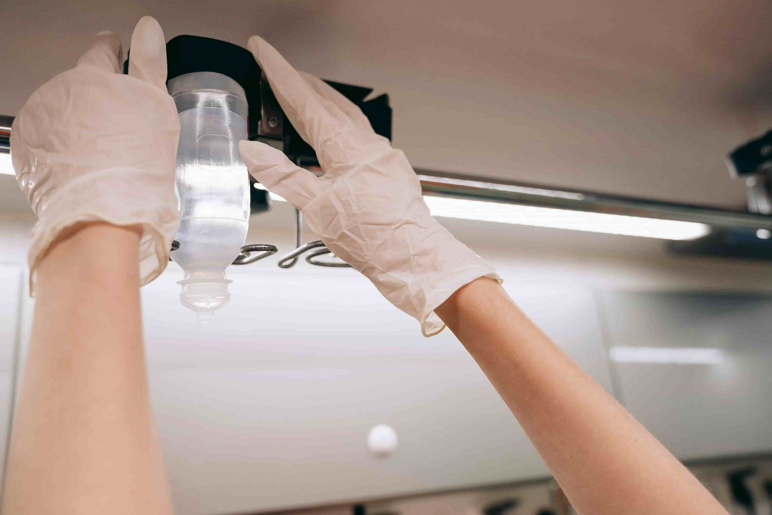 A person wearing white gloves is adjusting or hanging an IV fluid bag on a metal pole in a medical setting, raising the question: How Long Does a Mobile IV Treatment Take?.