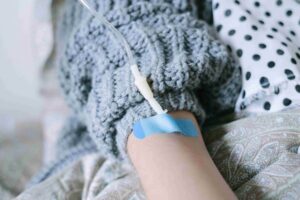 A person wearing a gray sweater has an IV line inserted into their hand, secured with blue medical tape, while lying on patterned bedding—raising questions like "Is Mobile IV Therapy Safe and Are There Any Risks?.