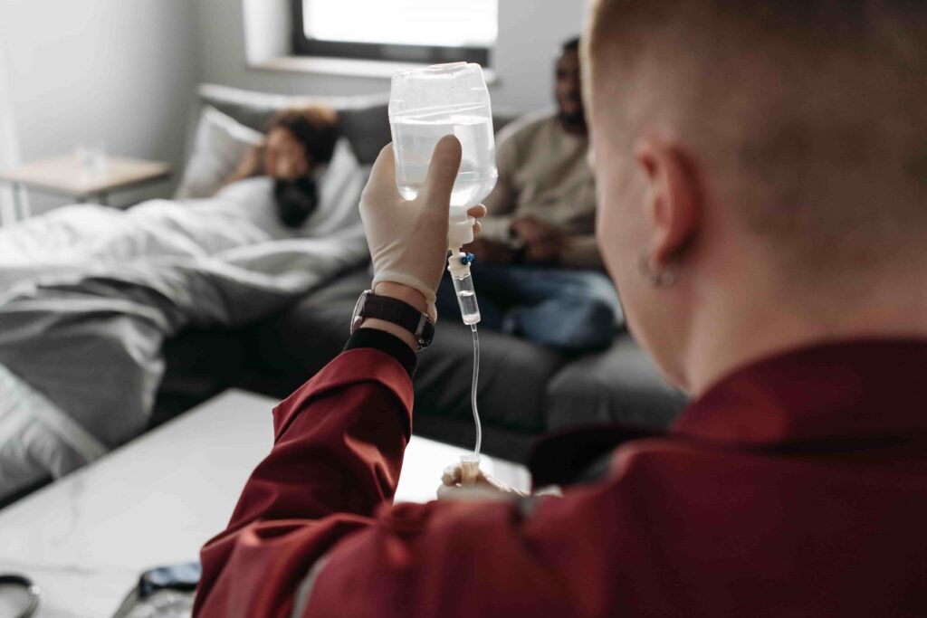 A medical professional prepares an IV drip while a patient lies in bed in the background and another person sits nearby, reflecting common scenes where people might wonder: Is Mobile IV Therapy Covered by Insurance in British Columbia?.