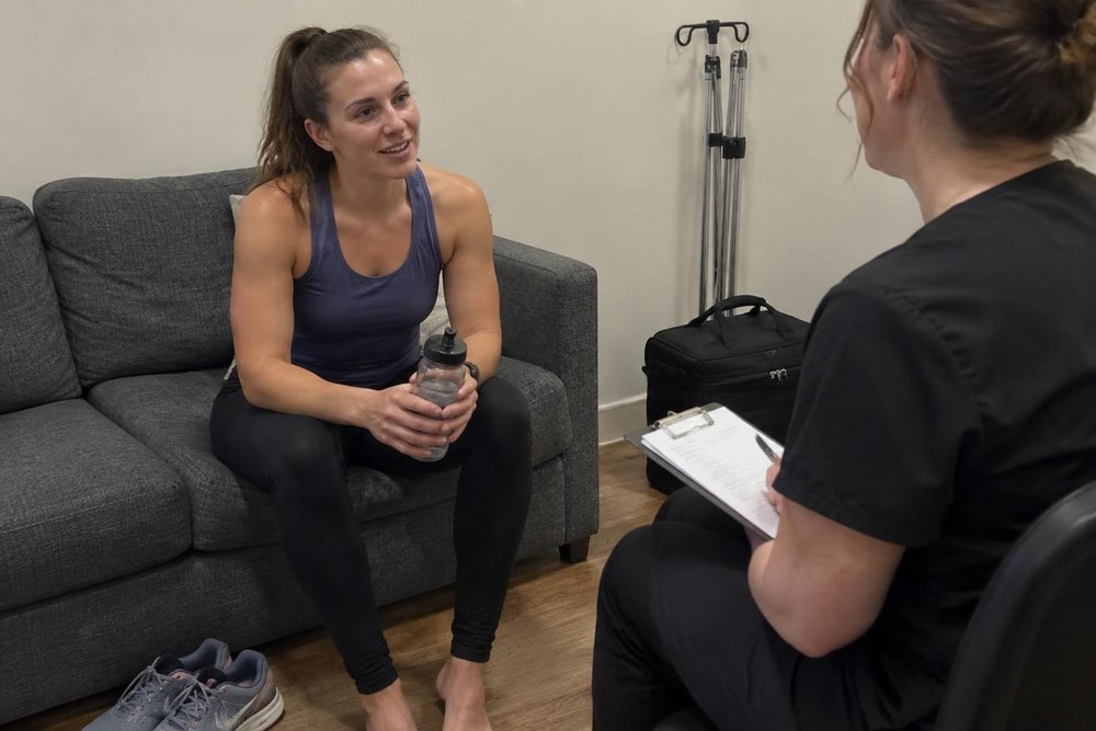A woman in athletic wear sits on a couch holding a water bottle, talking to another woman in scrubs who is holding a clipboard.